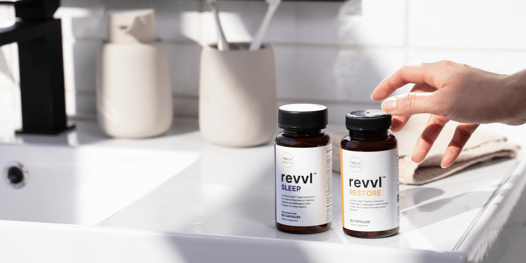 A person reaching for their bottles of Revvl Restore (for stress management) and Revvl Sleep (for melatonin-free sleep support) on the clean, white bathroom counter.  